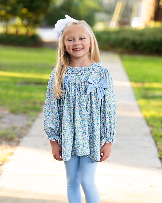 A young girl with blonde hair wearing a light blue floral long-sleeve blouse with large bows on the chest, light blue leggings, and a white hair bow, standing on a concrete path in a sunny outdoor setting.