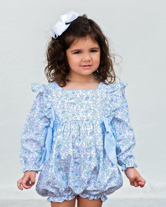 A young girl with curly brown hair wearing a light blue and white floral print long-sleeve romper with ruffled shoulders and a matching white bow headband, photographed against a plain light gray background.