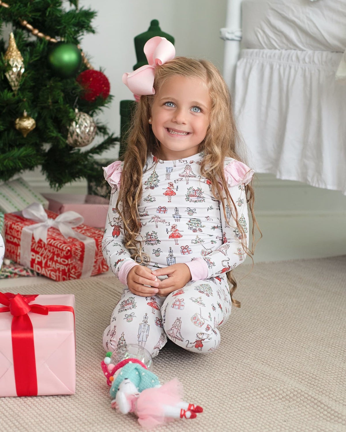 A young girl with long blonde hair sits on a carpeted floor in a cozy room decorated for Christmas. She is wearing a white long-sleeve pajama set with pink ruffled shoulders and a festive holiday print featuring Santa Claus, reindeer, and snowflakes.