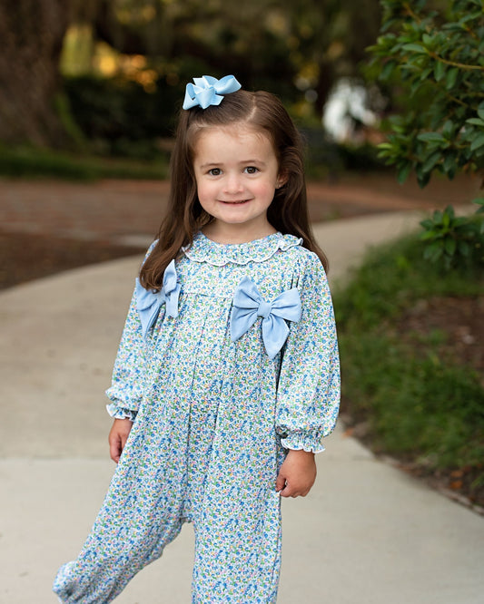 A young girl with long brown hair wearing a light blue floral jumpsuit with large bows on the chest and waist, standing on a concrete path outdoors.
