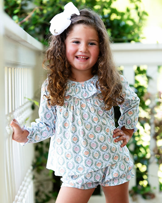A young girl with long curly brown hair wearing a matching long-sleeve top and shorts set in a light blue floral print, accessorized with a large white bow, posing outdoors on a porch.
