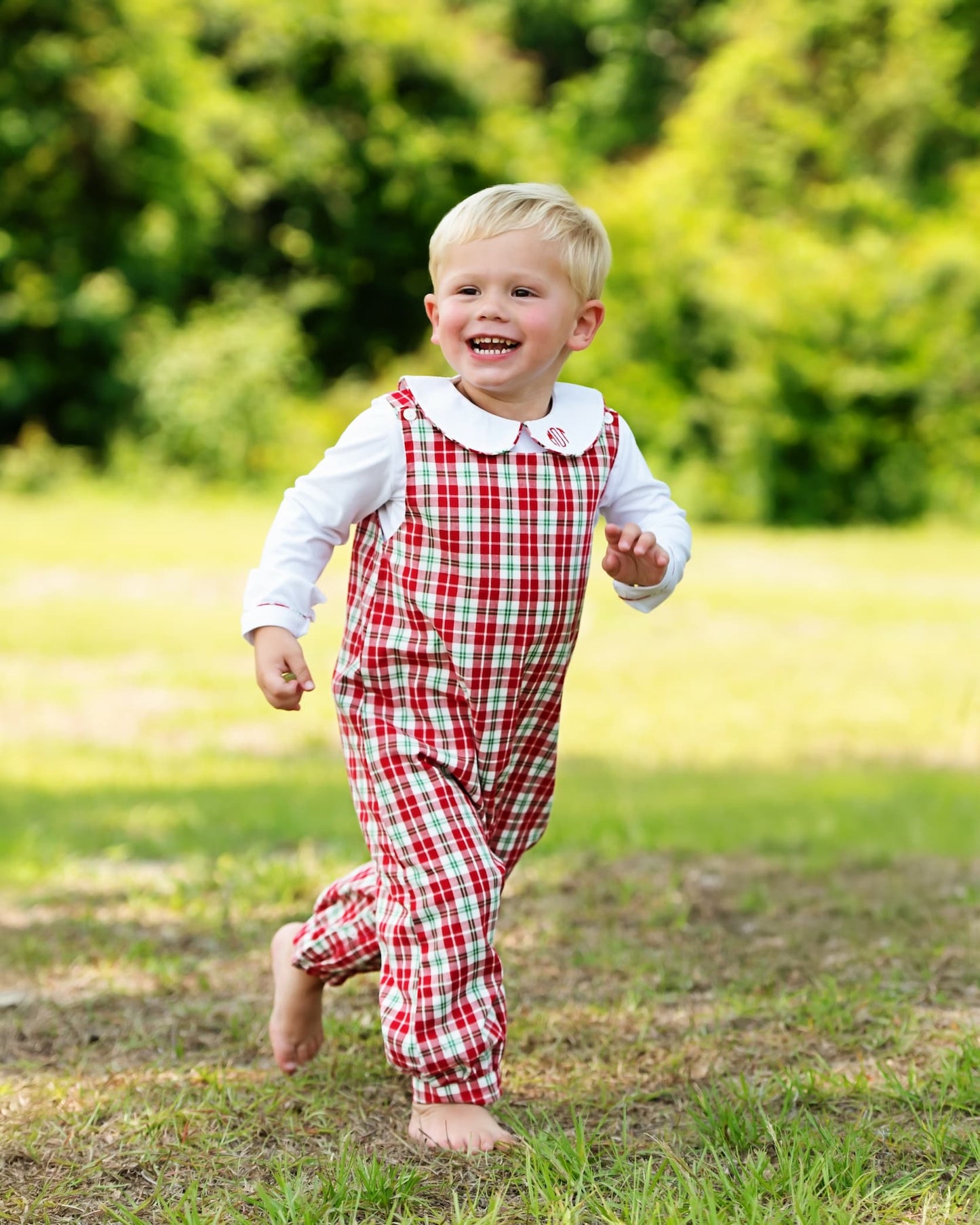 A young toddler boy running barefoot on a grassy lawn, wearing a red and white gingham check overall jumpsuit over a white long-sleeve shirt with a Peter Pan collar.