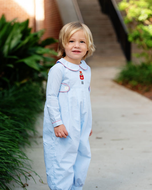 A young toddler with blonde hair stands on an outdoor concrete path wearing a light blue long-sleeve baby jumpsuit with white collar trim and a small red embroidered detail on the chest.