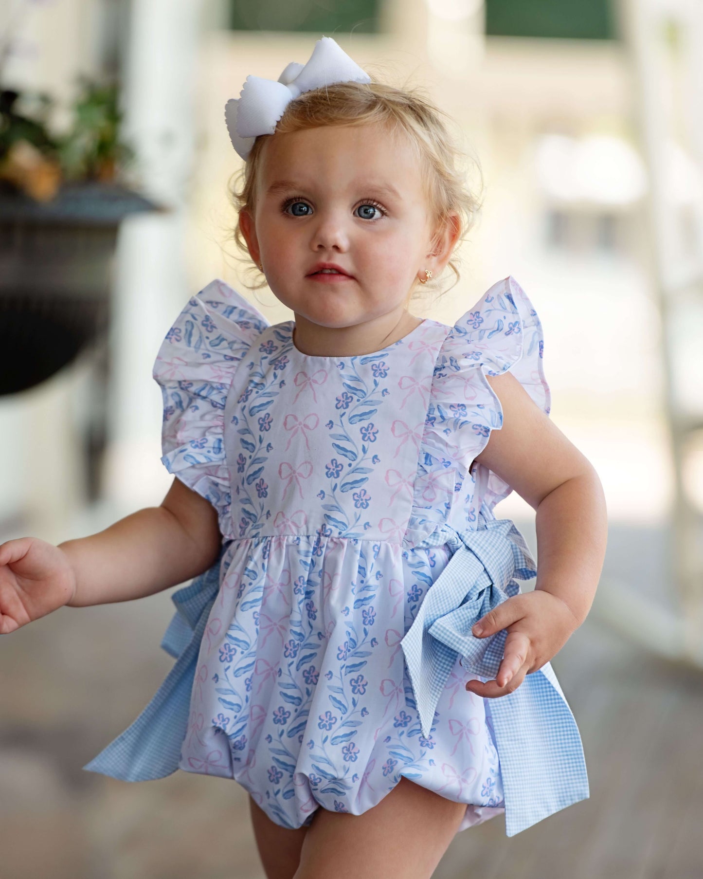 Baby girl in a floral romper with a bow, standing outdoors.