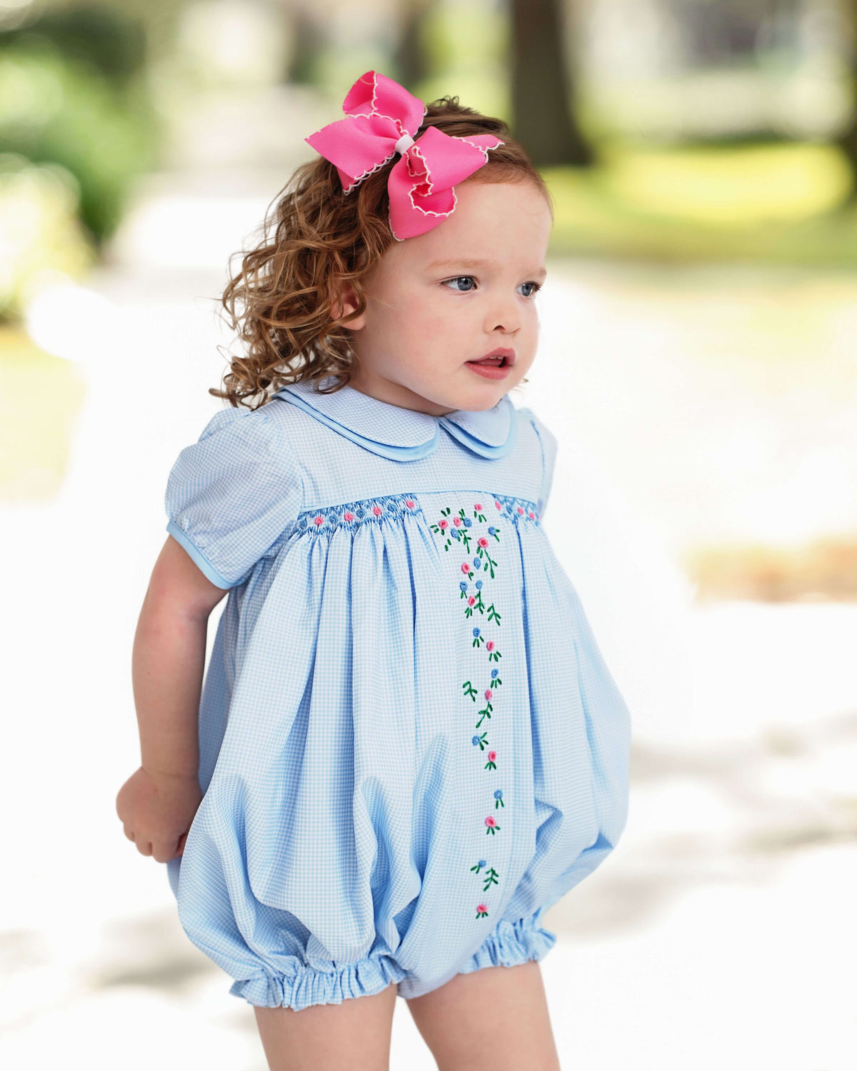 Baby girl wearing a light blue gingham bubble with floral embroidery outdoors.