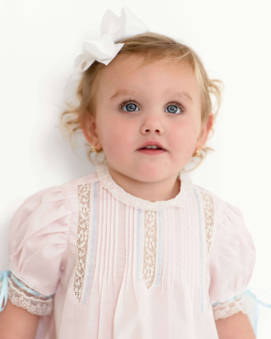 Baby girl wearing a pink dress with lace details and a white bow in her hair.