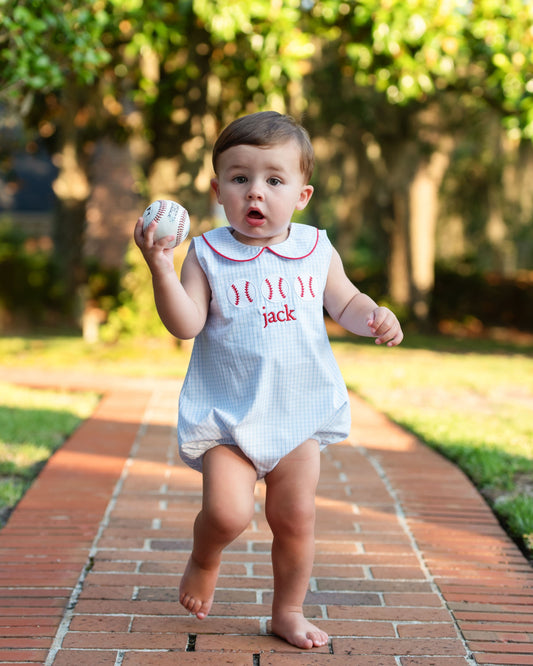 Baby in a white dress with red lettering holding a baseball on a brick path outdoors.
