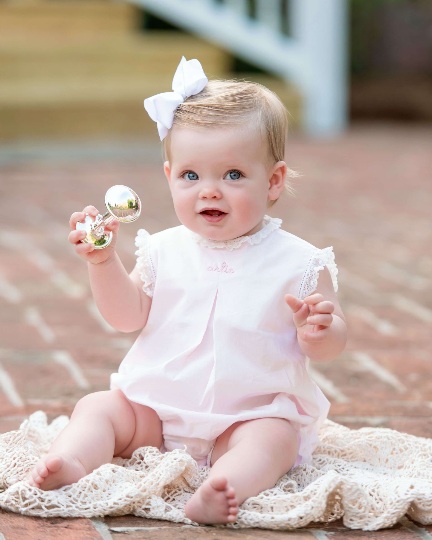 Baby in a white outfit with a bow, sitting on a textured surface.