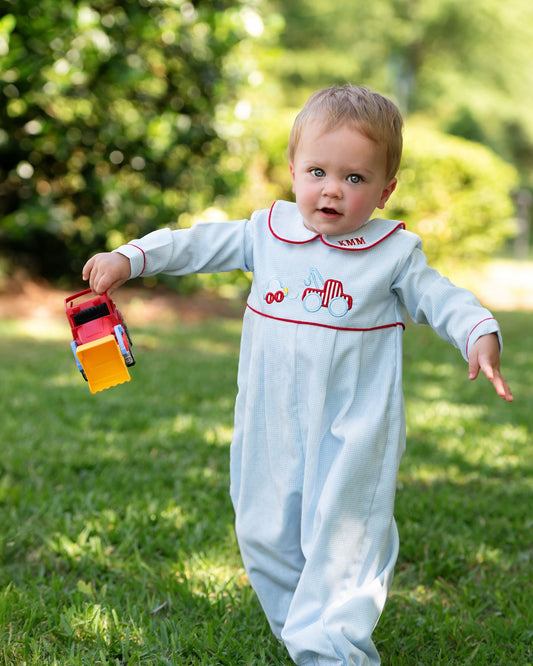 Baby in a white outfit with red trim holding a toy truck outdoors.