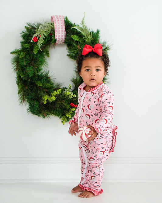 Baby in pink pajamas with a red bow standing in front of a Christmas wreath.