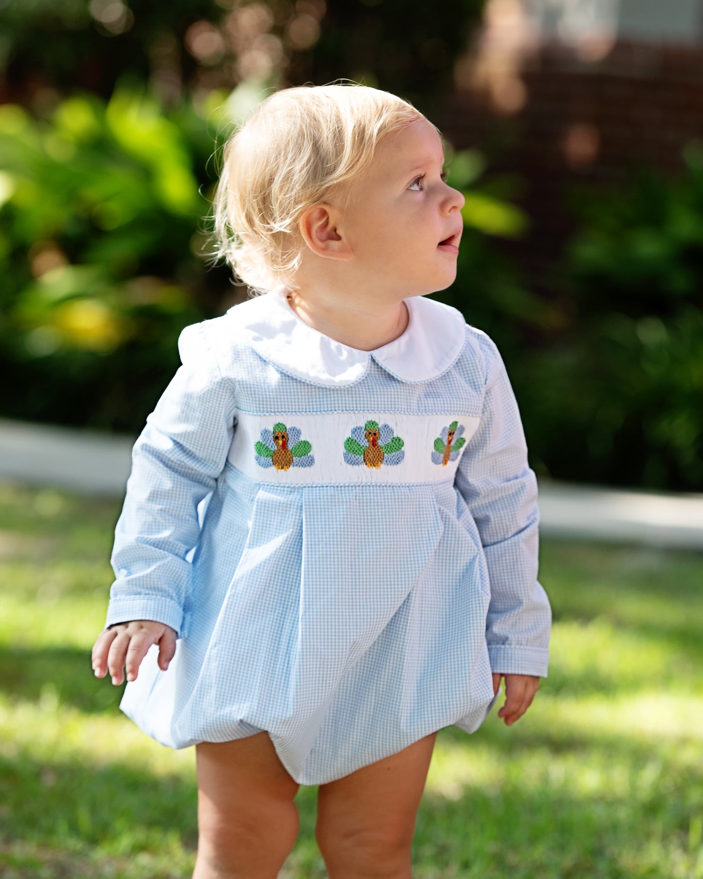 Baby wearing a light blue bubble with white collar and smocked turkeys outdoors.