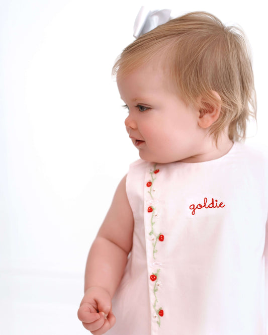 Baby wearing a pink dress with red buttons and the name 'goldie' on a white background