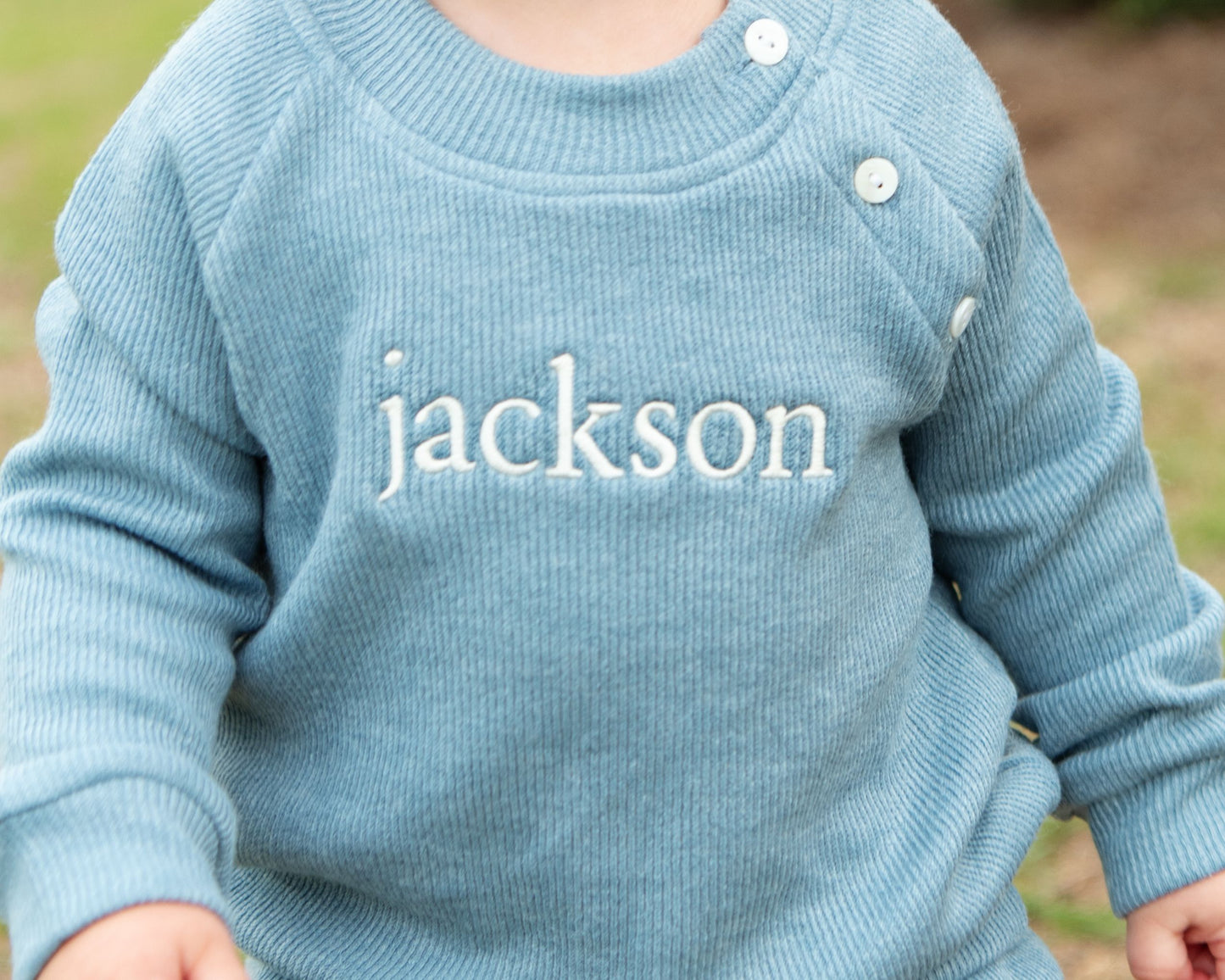 Blue sweater with 'jackson' embroidered on it against a blurred natural background