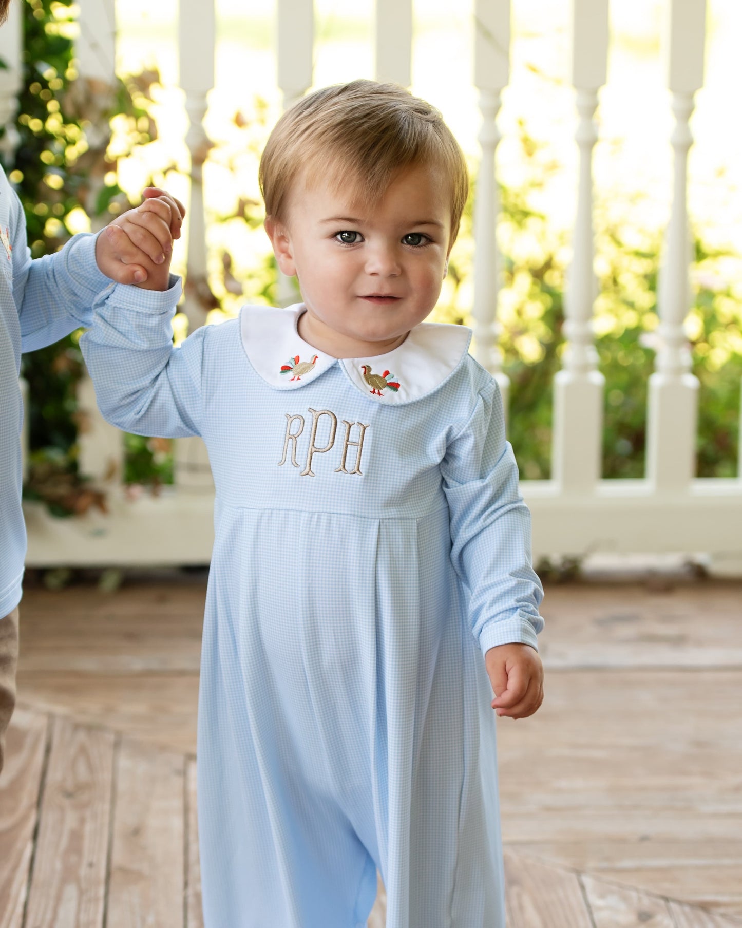 Boy in a blue gingham long romper with embroidered turkeys on collar with monogram "rPh" standing on a wooden floor.