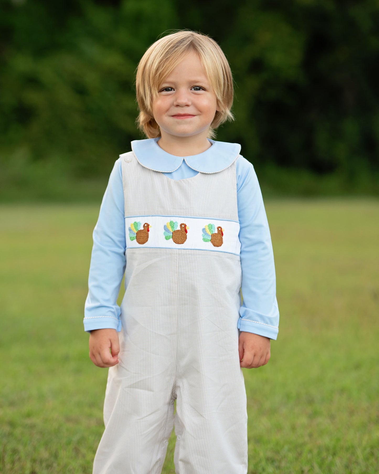 Boy wearing a blue and khaki long romper with smocked turkeys in an outdoor setting.