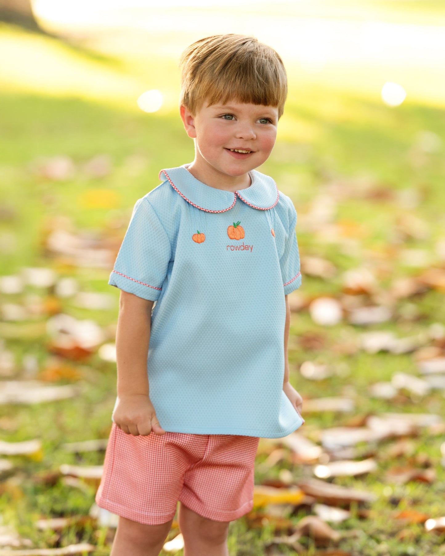 Boy wearing a light blue shirt with pumpkin designs and orange shorts standing in a grassy area.