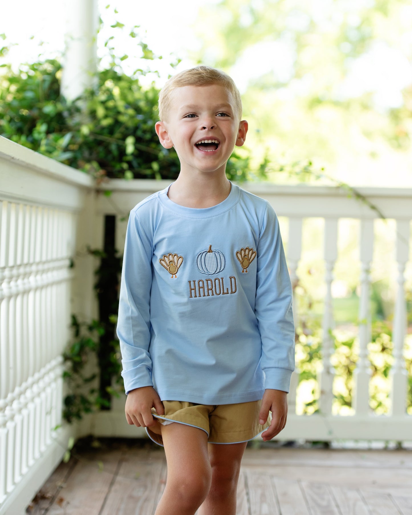 Boy wearing a turkey and pumpkin blue and khaki short set with 'Harold' monogram, standing on a porch.