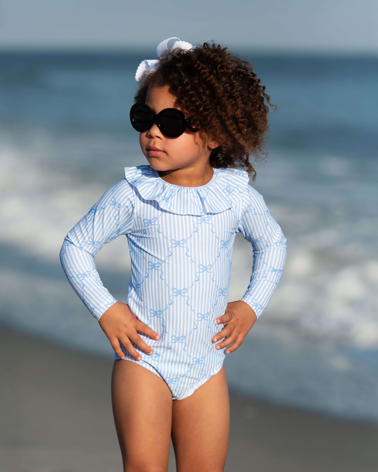 Child in a blue striped swimsuit and sunglasses standing on a beach with ocean in the background