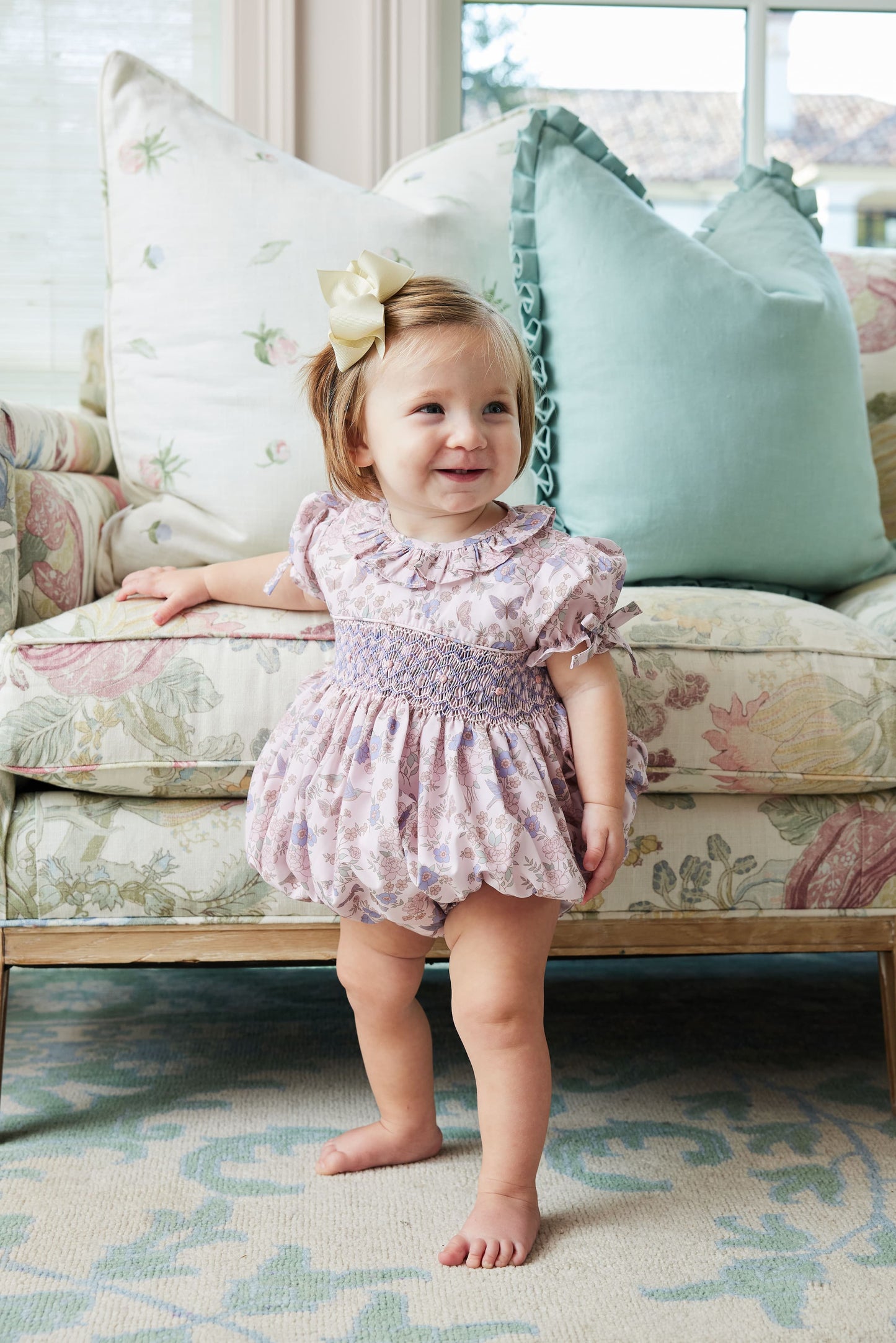 Child in a floral dress standing in a living room with floral-patterned sofa and cushions.