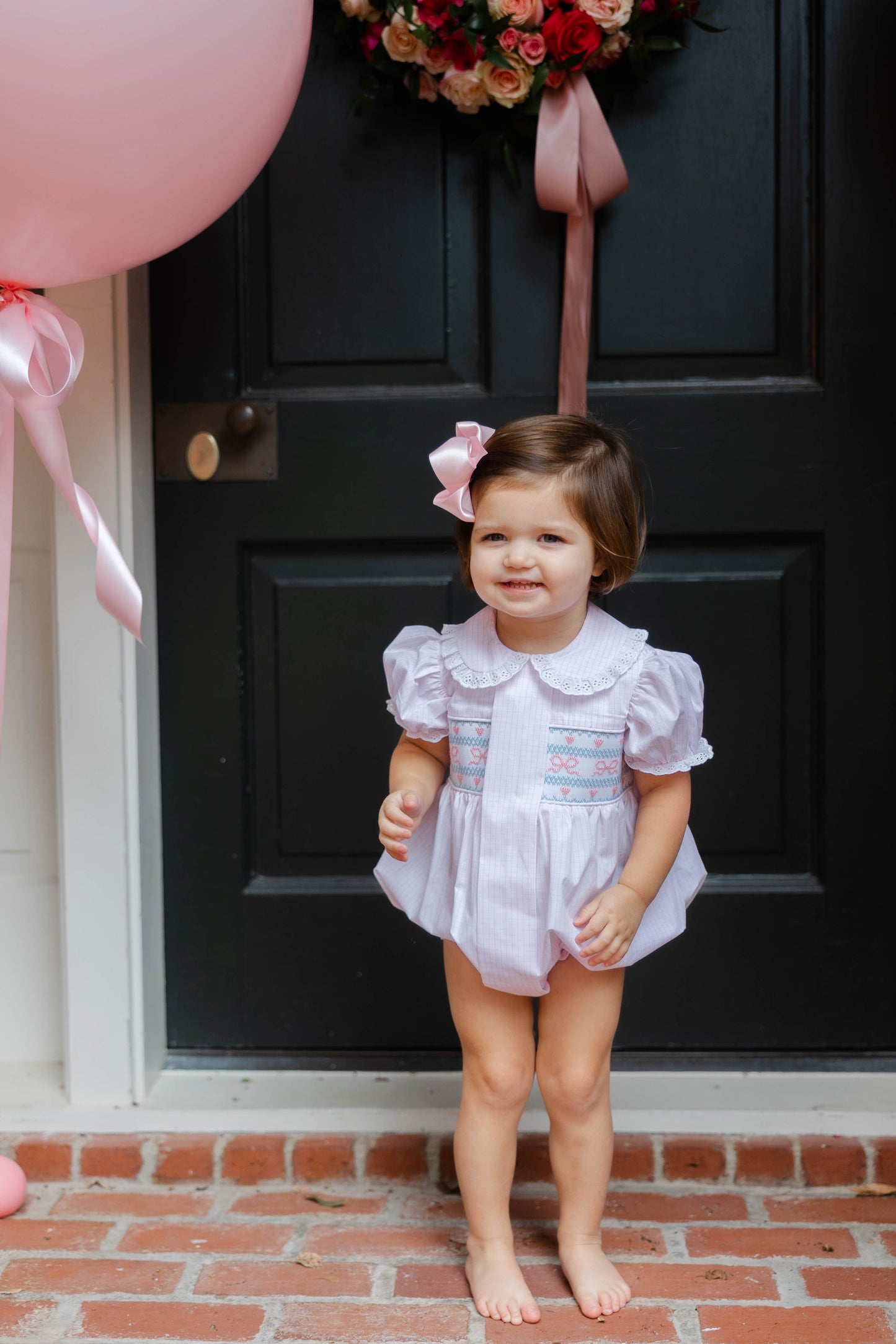 Child in a white dress with pink bow standing in front of a black door with pink balloons.