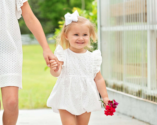 Child in a white eyelet bubble holding hands with a little girl, standing outdoors.