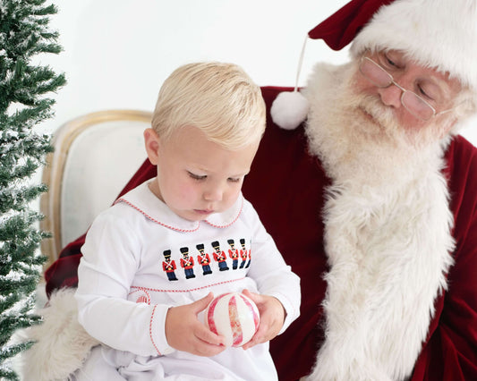 Child in a white outfit with embroidered Christmas nutcrackers sitting next to Santa Clause