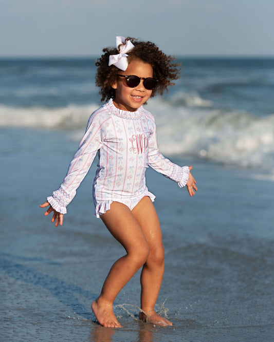 Child in a white swimsuit and sunglasses standing on a beach with ocean waves in the background