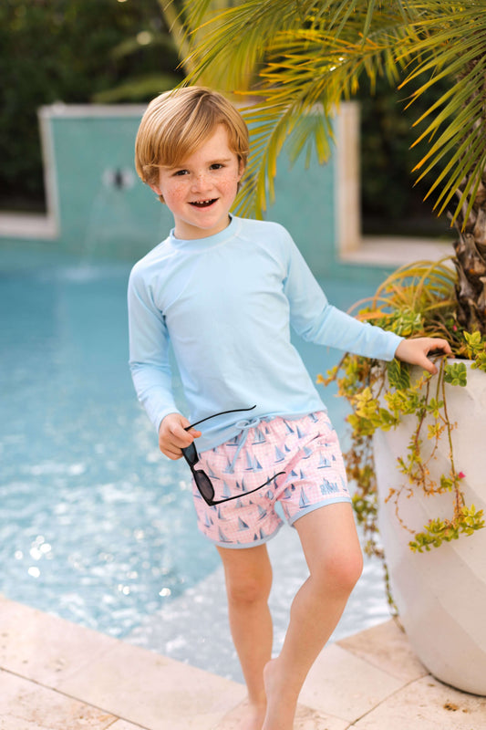 Child in light blue long-sleeve shirt and pink shorts standing by a pool with sunglasses.