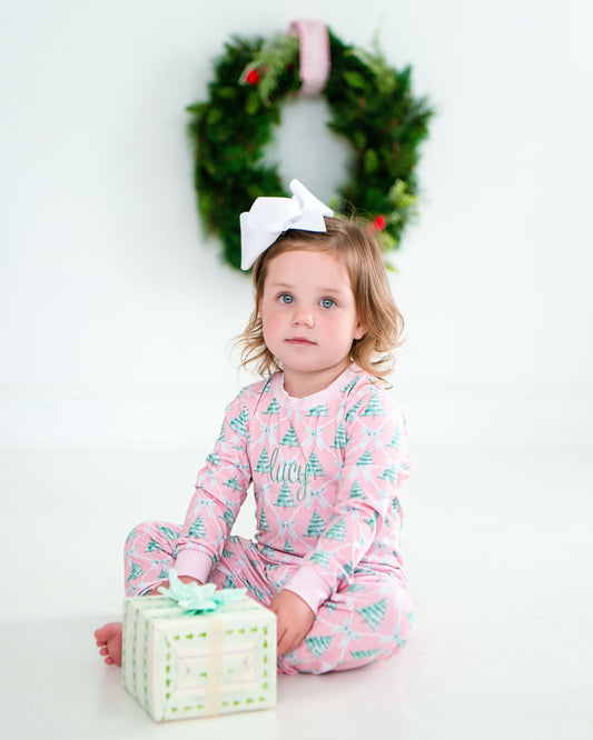 Child in pink pajamas with Christmas tree pattern, sitting next to a gift box with a wreath in the background.