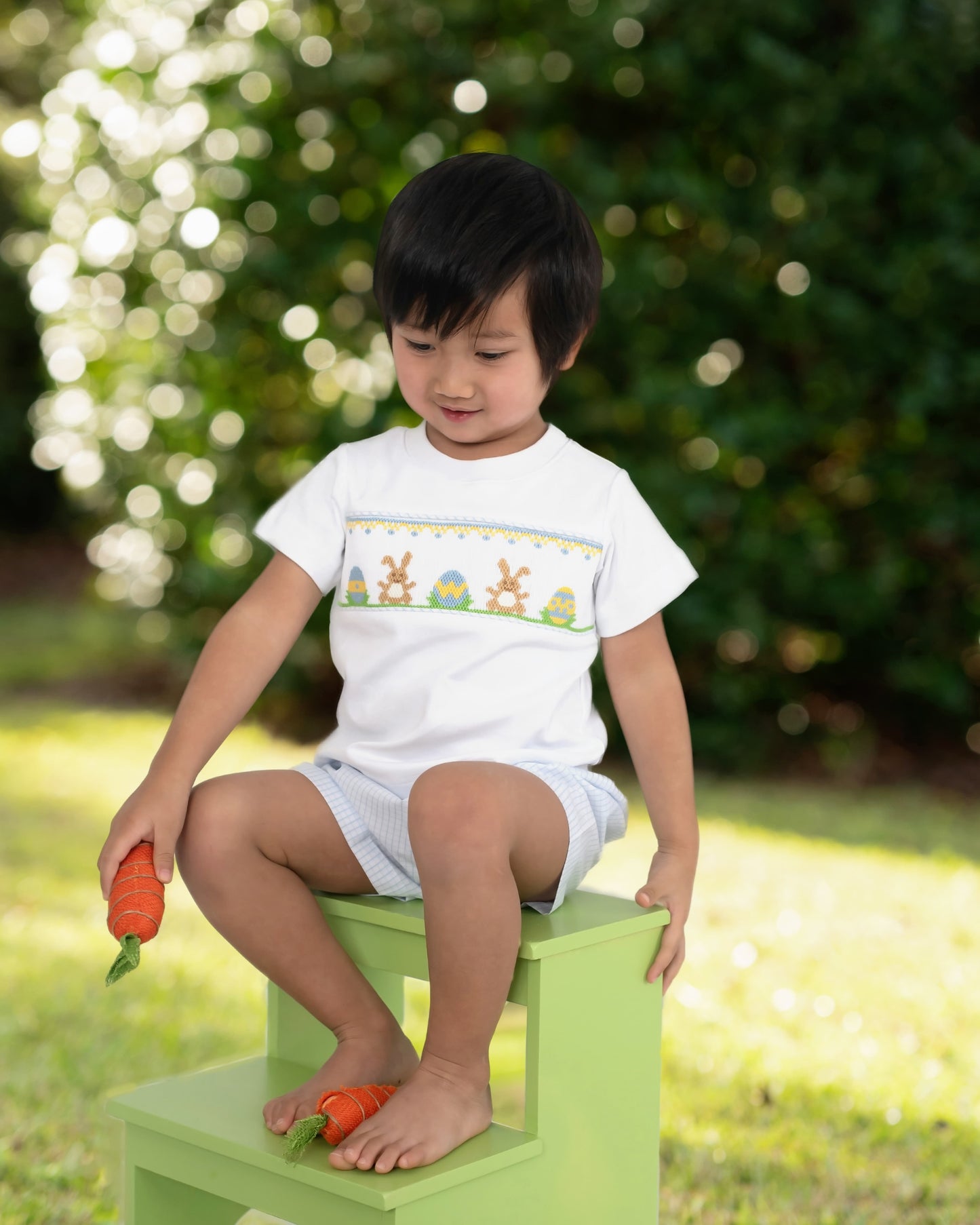 Child sitting on a green step stool holding carrots, wearing a white shirt with bunny designs outdoors.