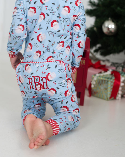 Child wearing a blue Christmas-themed onesie with Santa Claus pattern and monogram 'RBB' in front of a decorated Christmas tree.