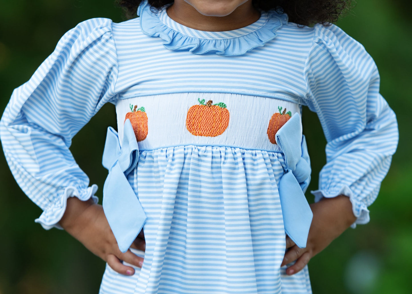 Child wearing a blue and white striped diaper set with pumpkin embroidery, standing against a blurred green background.