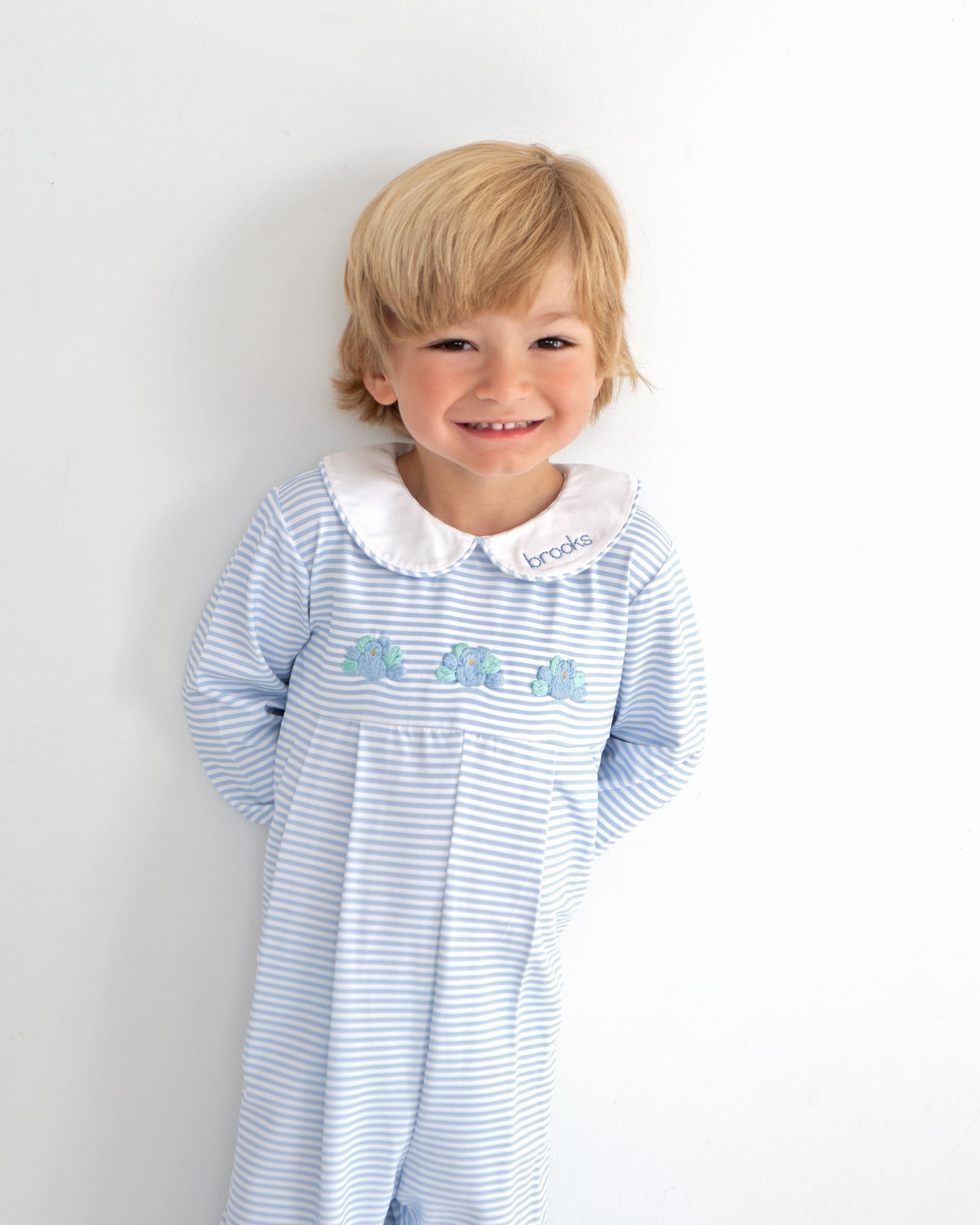 Child wearing a blue and white striped long romper with a white collar and embroidered turkeys against a plain background