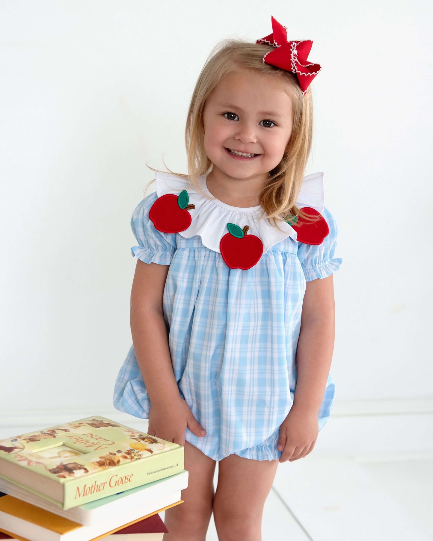 Child wearing a blue checkered outfit with red apple appliqués on a white background