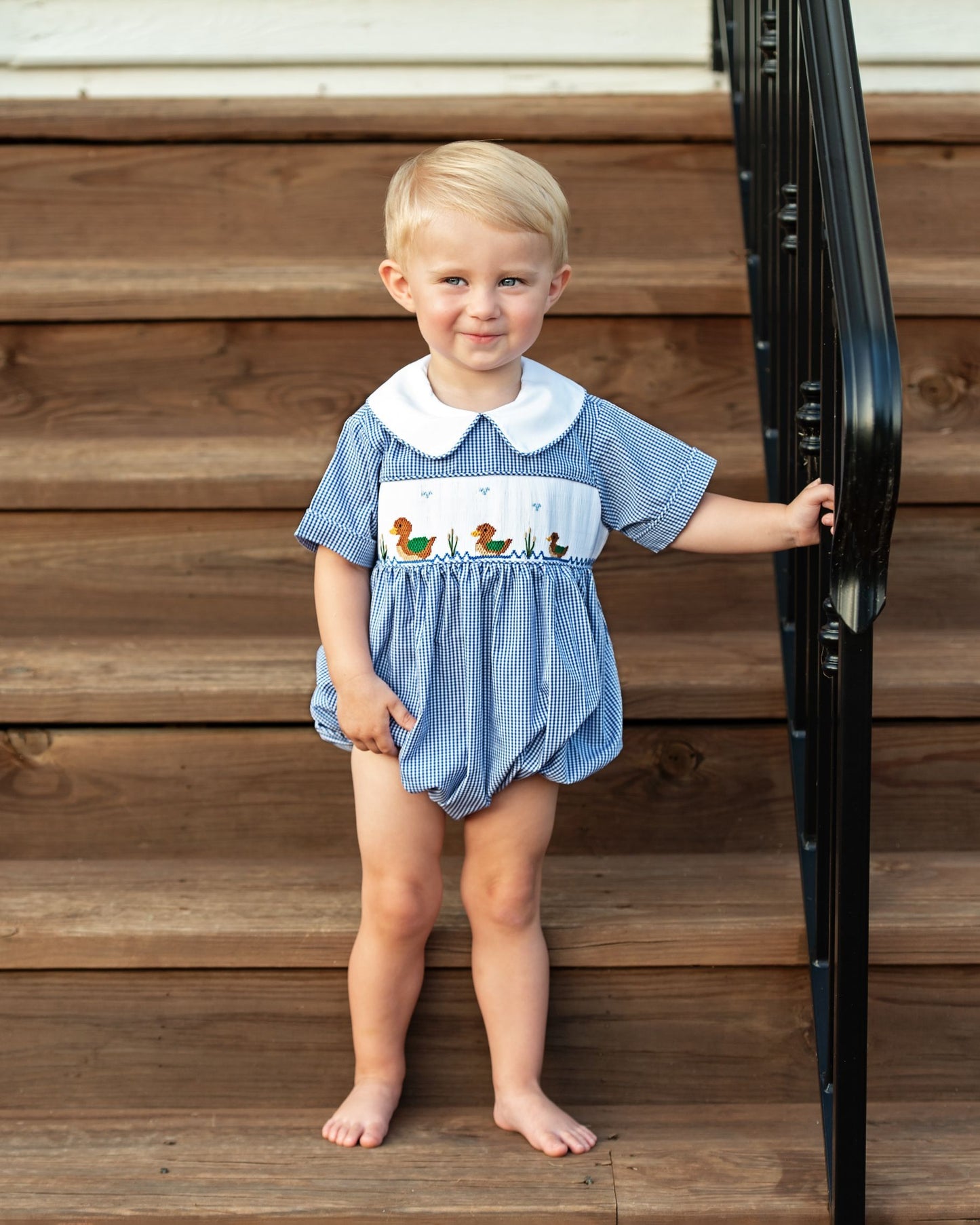 Child wearing a blue checkered romper with a collar and duck design, standing on wooden steps.