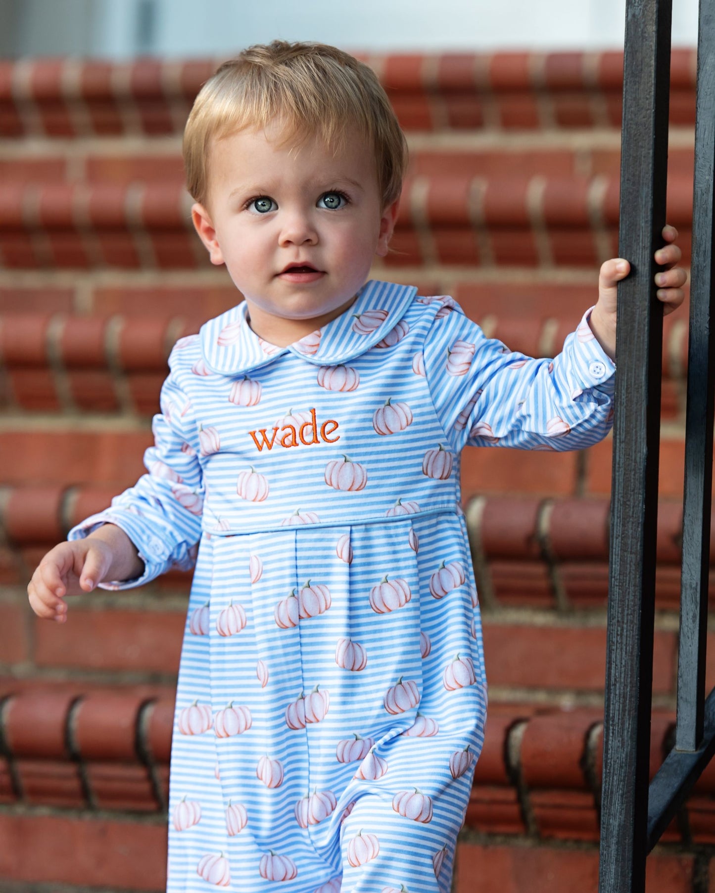 Child wearing a blue onesie with orange text and pumpkin pattern, standing on red brick steps.