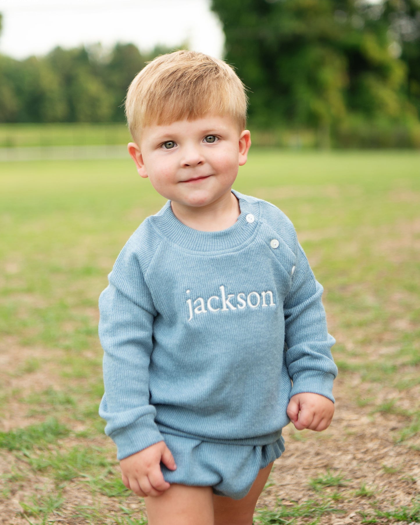 Child wearing a blue outfit with 'jackson' printed on it, standing in a grassy field.