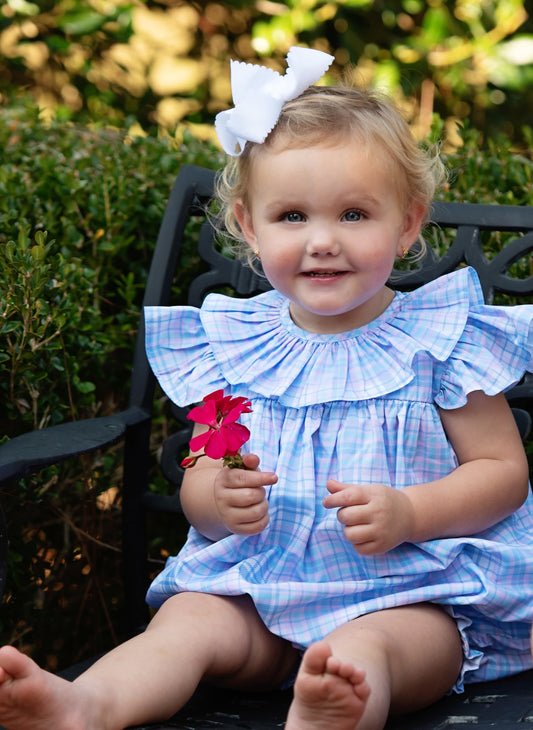 Child wearing a blue plaid bubble with a large white bow in hair, sitting outdoors.