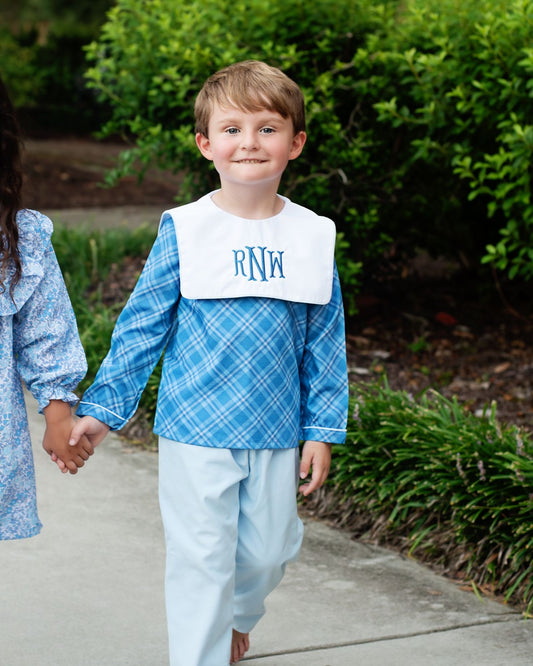 Child wearing a blue plaid shirt with a bib holding hands with another child outdoors.