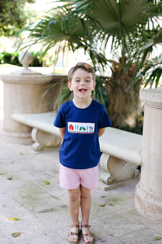 Child wearing a blue shirt with a graphic design, standing outdoors with greenery in the background
