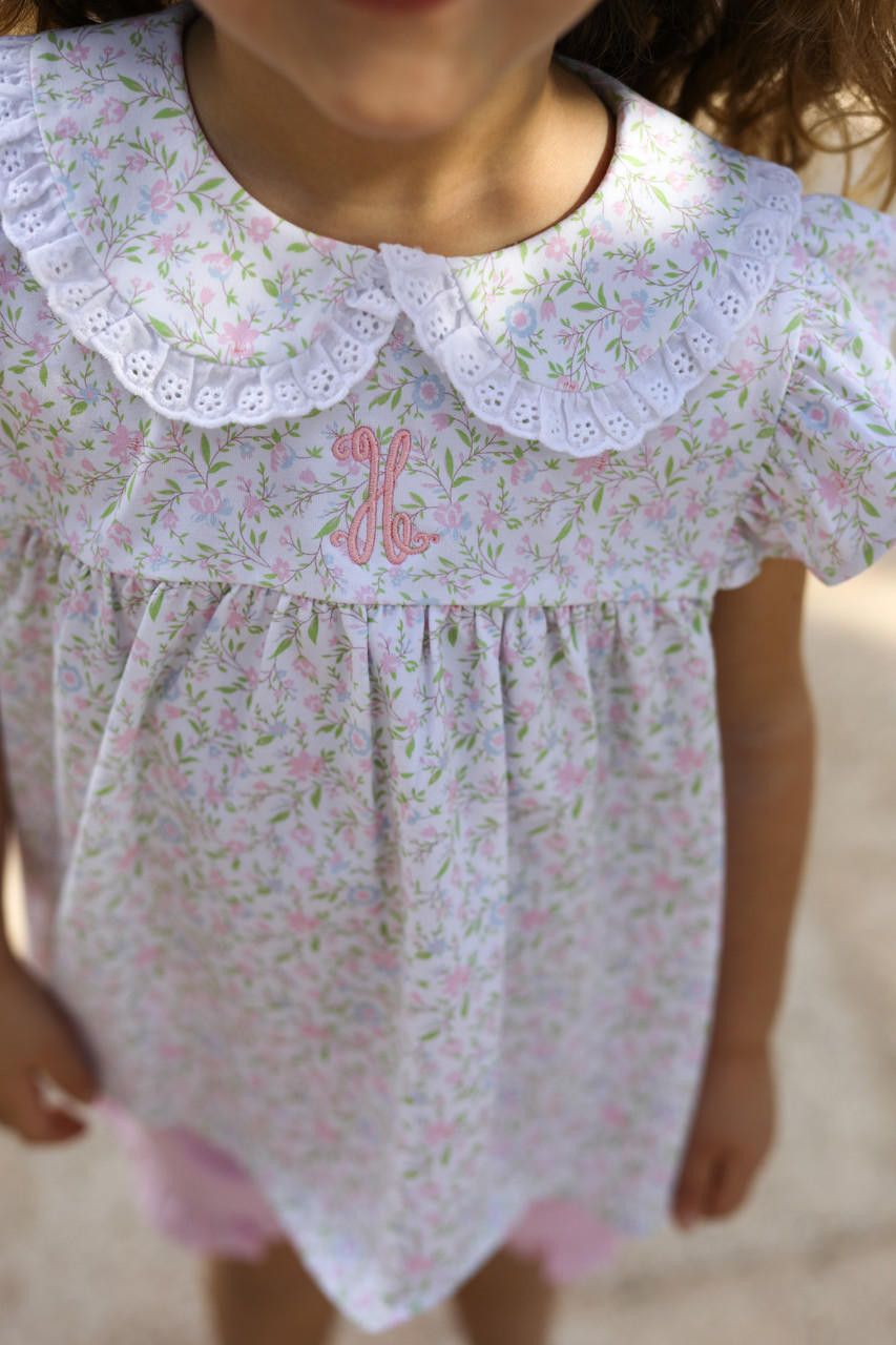 Child wearing a floral dress with a lace collar and pink letter 'H' on a blurred background