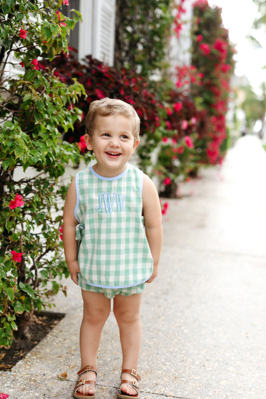 Child wearing a green checkered outfit standing on a sidewalk with flowers in the background