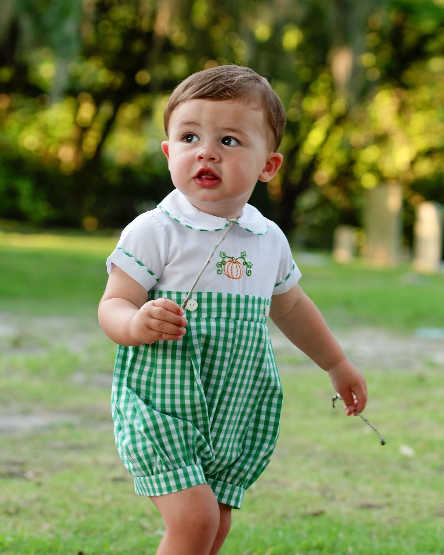 Young boy wearing a green checkered outfit with a white shirt featuring pumpkin designs, standing outdoors.