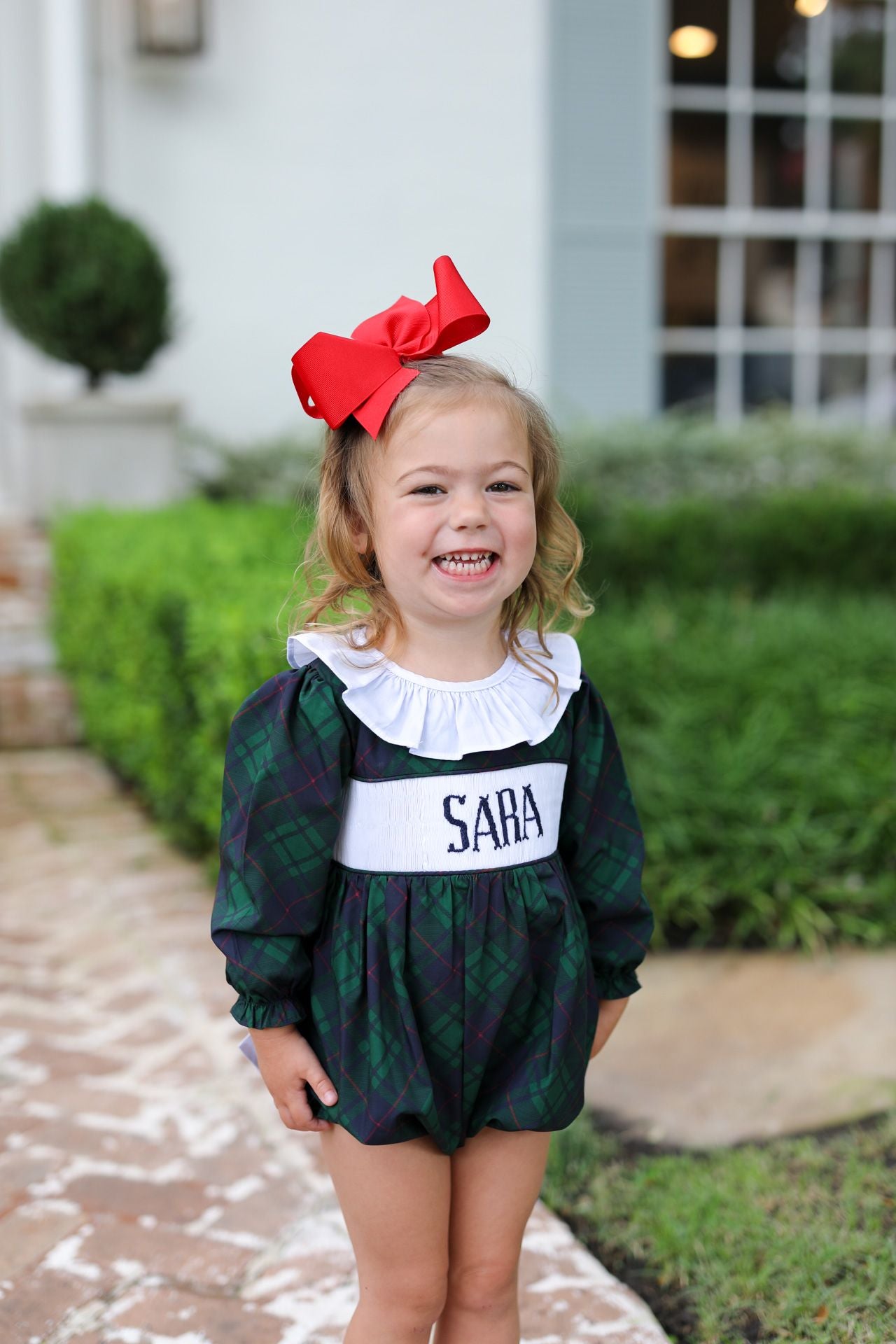 Child wearing a green plaid outfit with a red bow, standing outdoors.