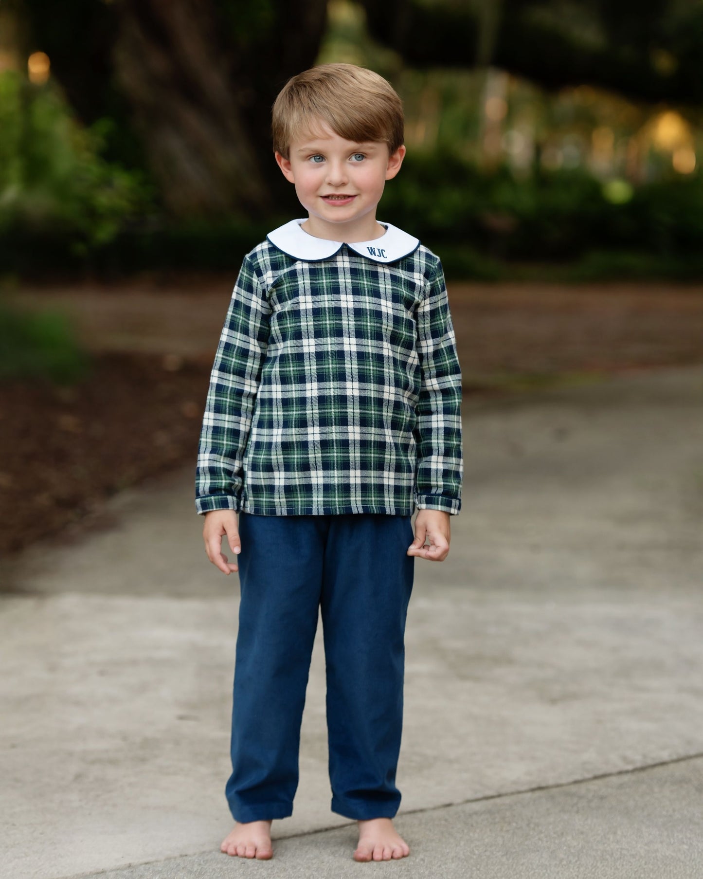 Child wearing a green plaid shirt with a white collar and blue pants standing on a sidewalk.