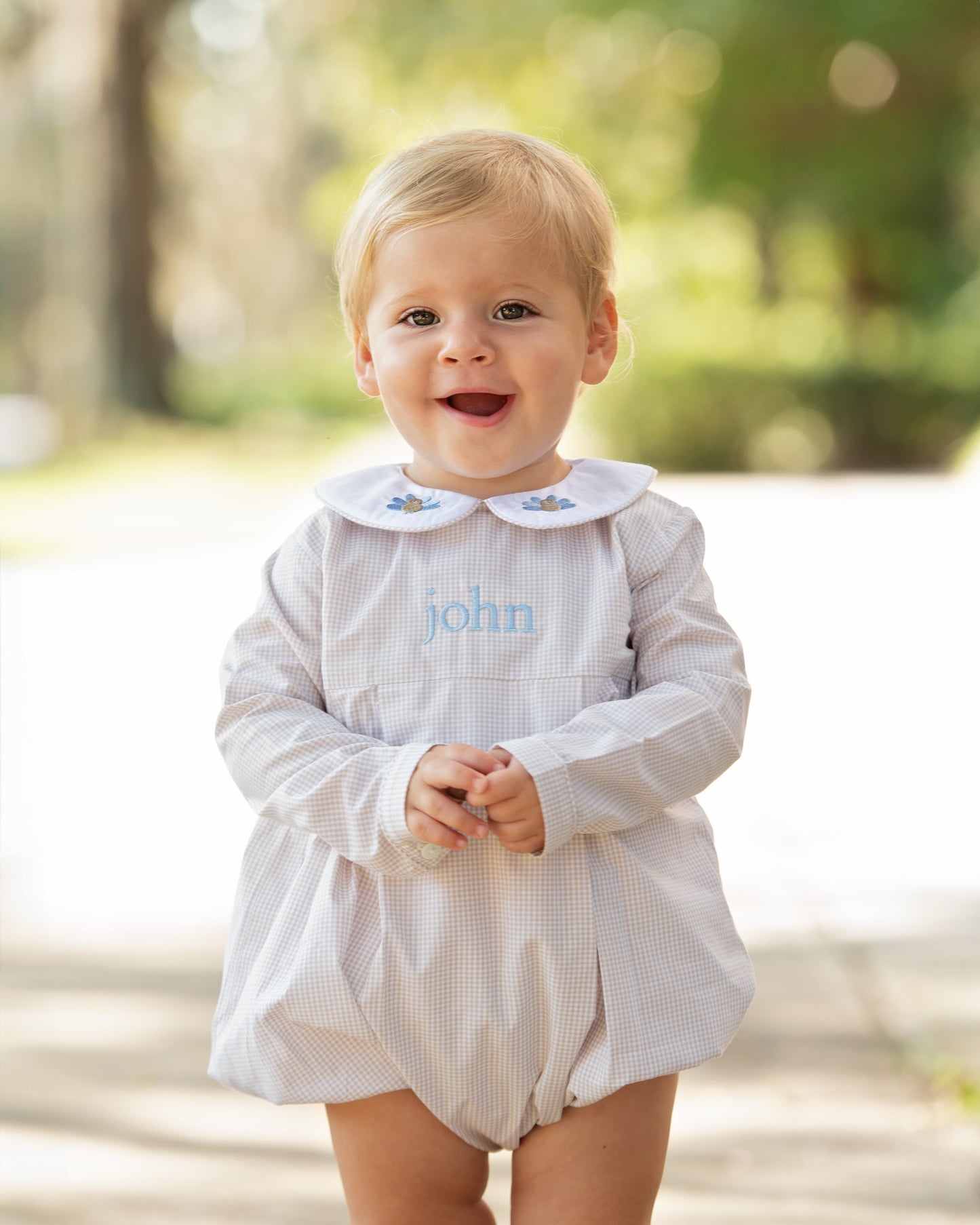 Child wearing a khaki gingham outfit with embroidered turkeys and a monogram on it, standing outdoors.