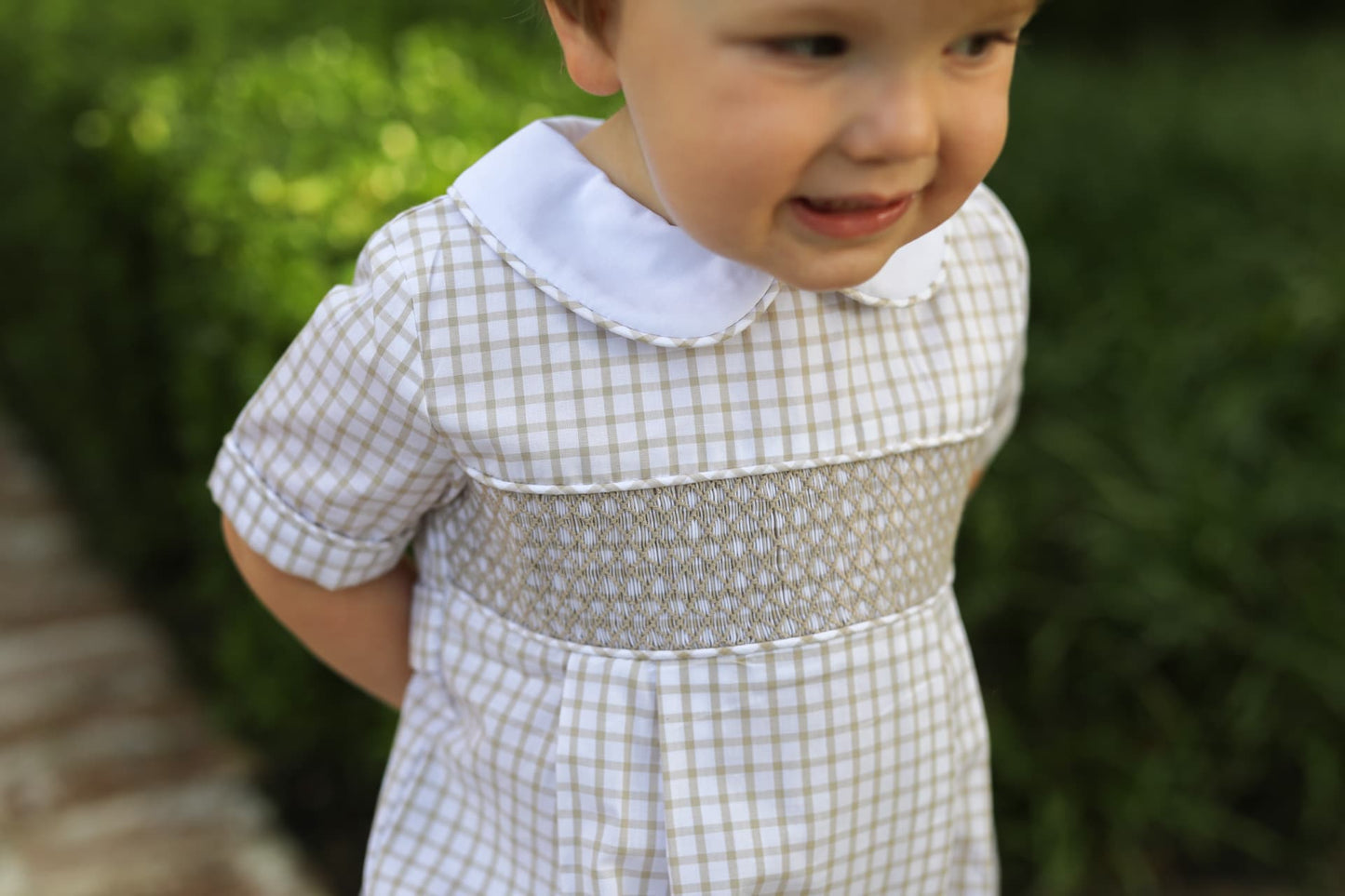 Child wearing a khaki smocked bubble with a white collar outdoors.