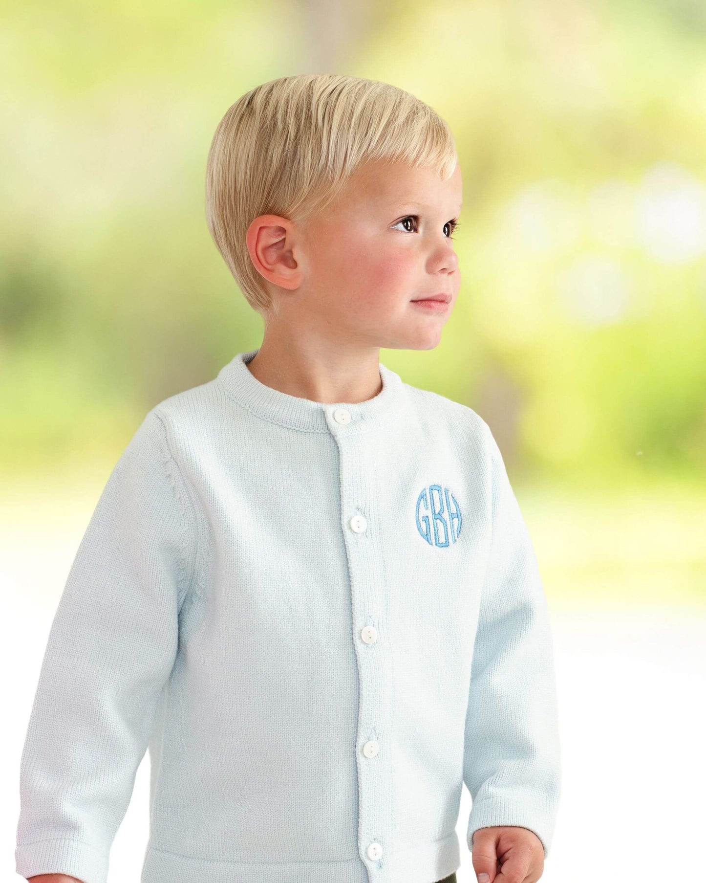Child wearing a light blue cardigan with monogram letters against a blurred natural background