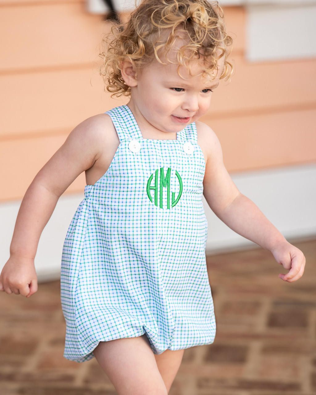 Child wearing a light blue checkered dress with green monogram on a wooden floor.
