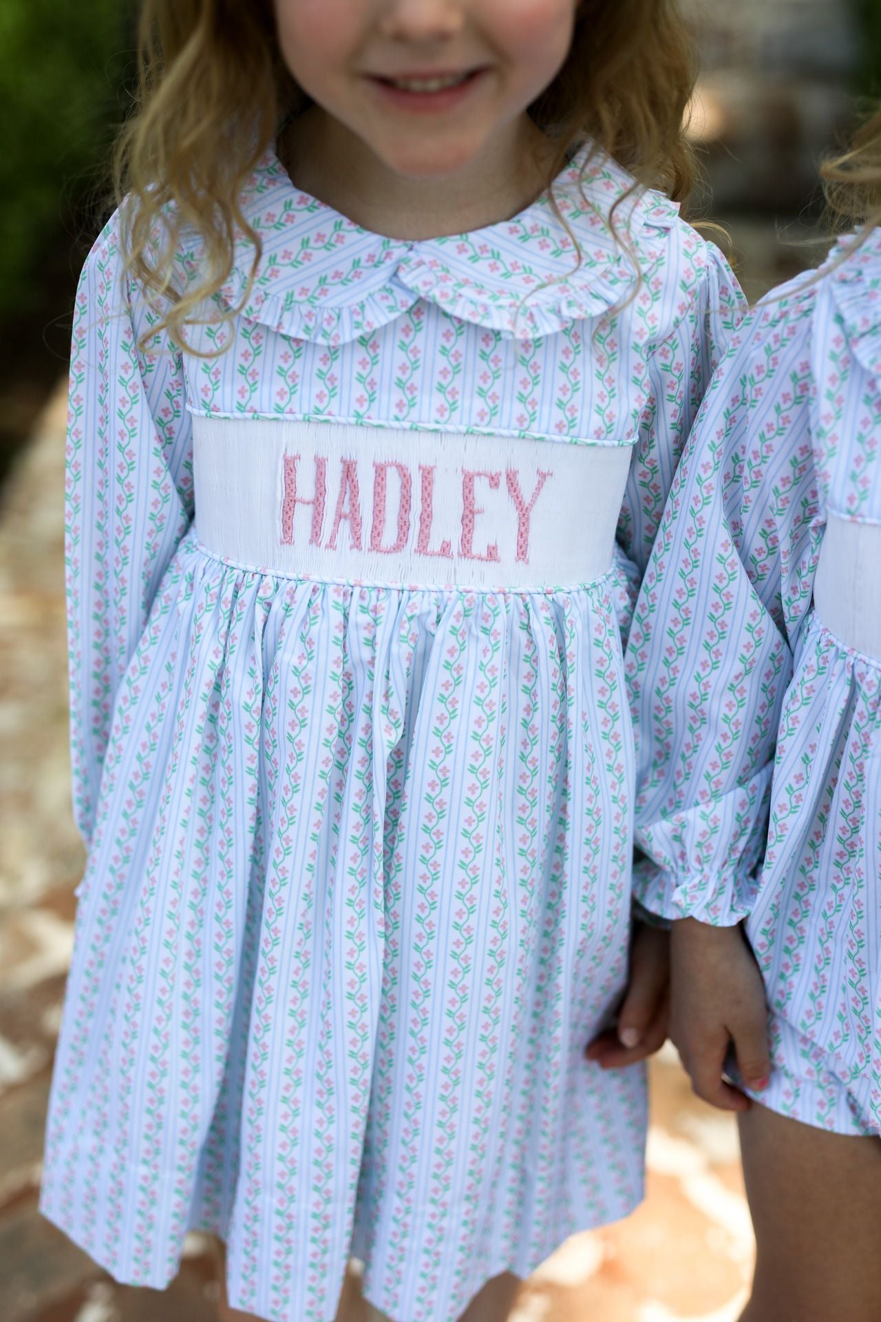 Child wearing a light blue dress with 'HADLEY' embroidered on it, standing outdoors.